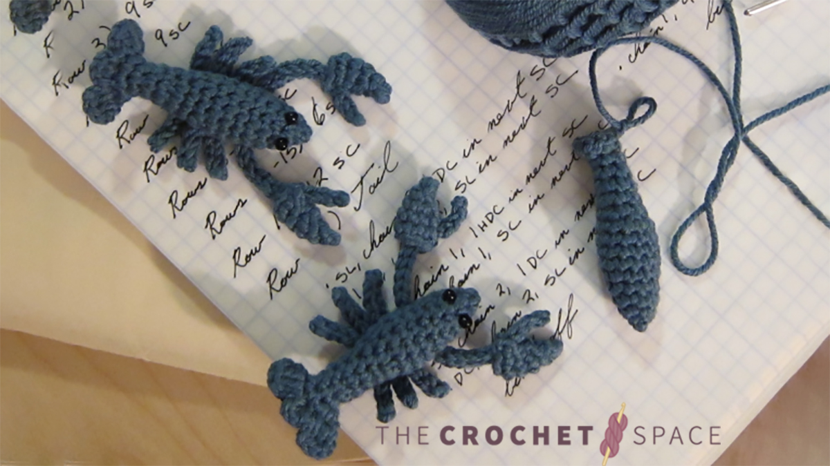 Little Leo Crochet Lobster. Two complete little lobsters, and one more body just started. Crafted in Blue yarn and laid out on an open white page in a book with writing on it || thecrochetspace.com