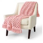 Love Net Crochet Blanket thrown over a chair. Dusky pink, love knot stitch with added tassels || thecrochetspace.com